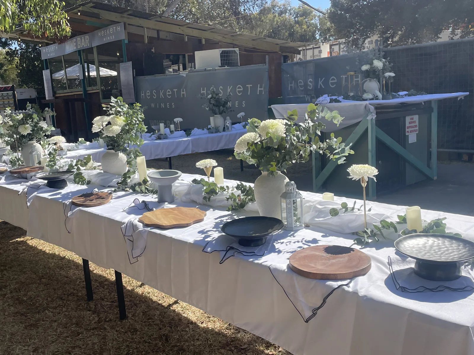 A long outdoor event table in white linen, with eucalyptus garland, white chrysanthemums, candles and timber boards