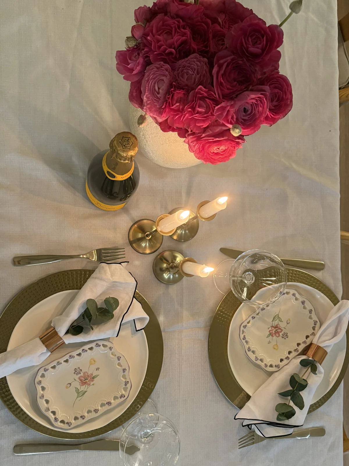 From above: a table for two set with gold chargers, floral side plates, linen napkins in copper rings with eucalyptus, brass candlesticks and pink ranunculus