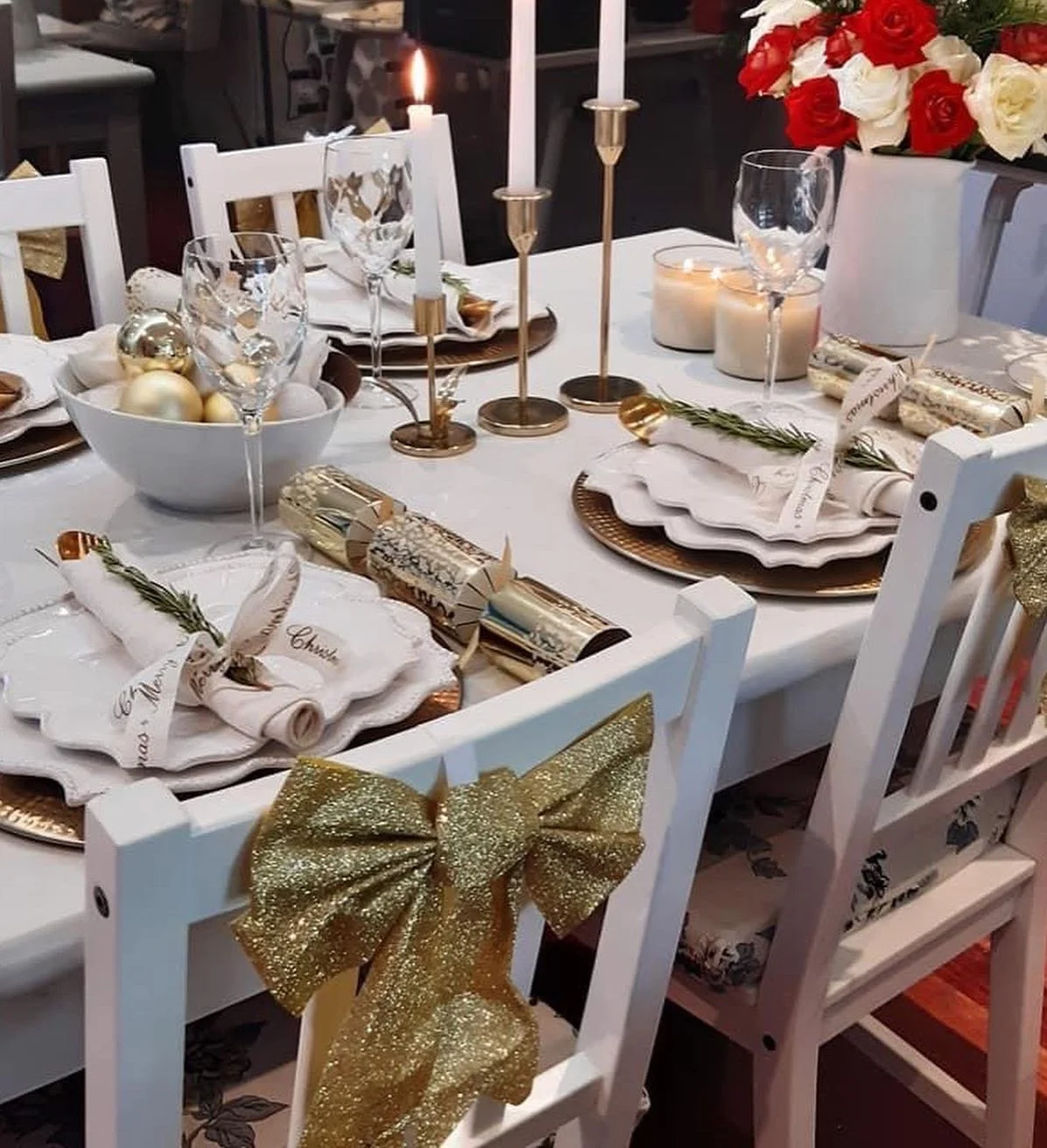 A Christmas table: lit white tapers in brass holders, red and white roses, gold chargers, rosemary-tied napkins, gold crackers and glitter bows on the chairs